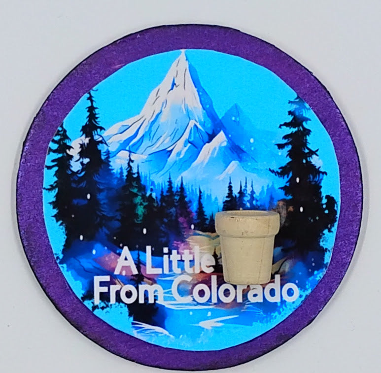 Handcrafted circlular purple magnet with a beautiful blue sky, snow covered mountains and a tiny little pot.