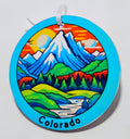 Handcrafted blue mountain ornament with a red sky, a green field and snow covered mountains. Says Colorado on the bottom. 