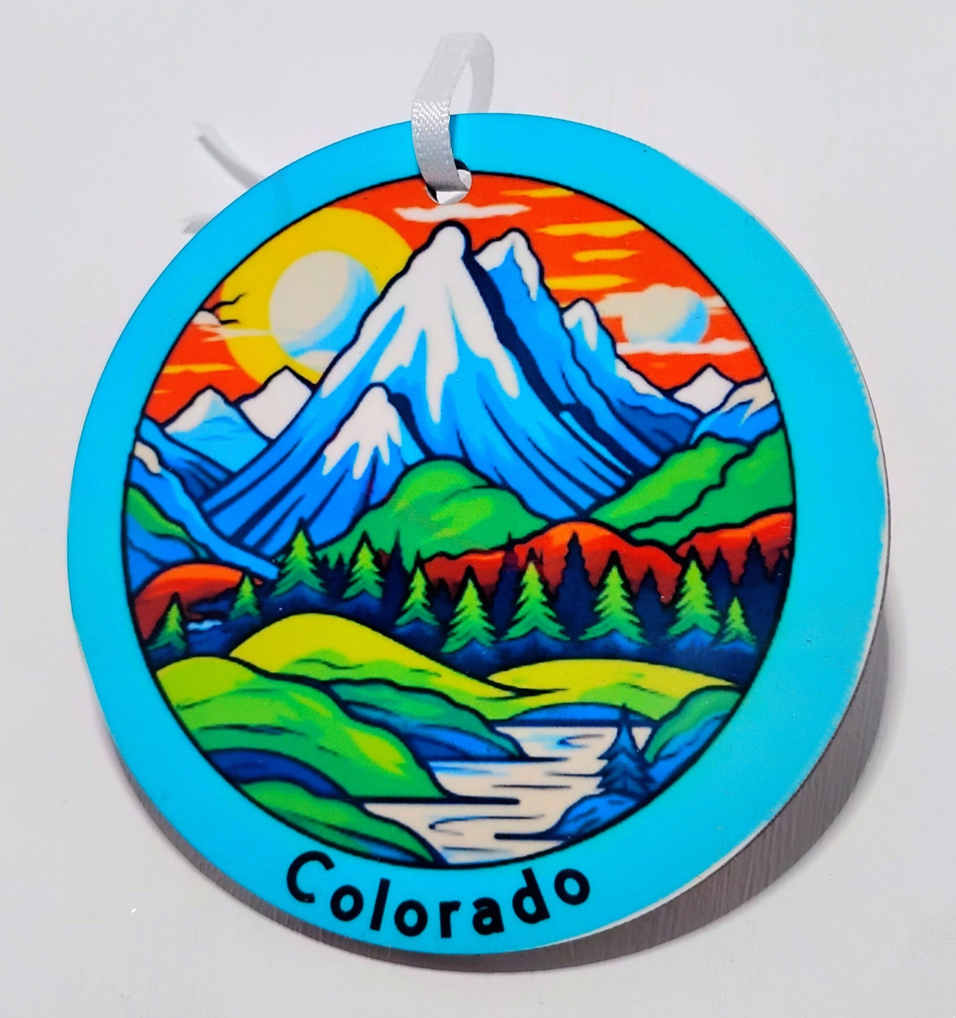 Handcrafted blue mountain ornament with a red sky, a green field and snow covered mountains. Says Colorado on the bottom. 