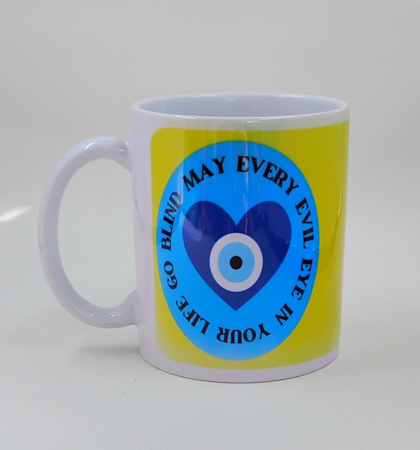 A coffee mug with an evil eye heart and the saying "may every evil eye in your life go blind" with a yellow background.