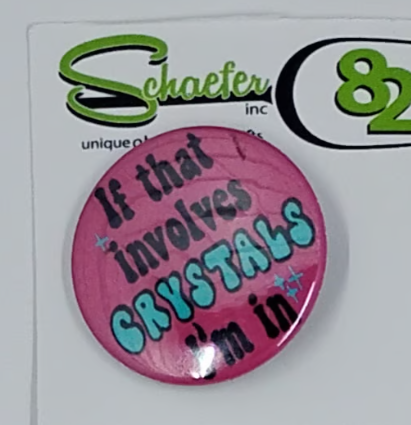 A small round pink hat pin with the saying "if that involves crystals, I'm in"