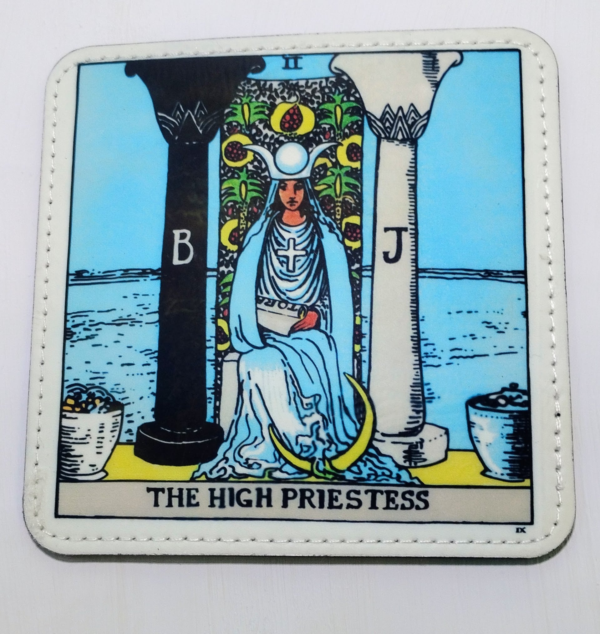 White pleather coaster that has the image from the tarot card "The high Priestess:, which has a woman sitting between two pillars. 
