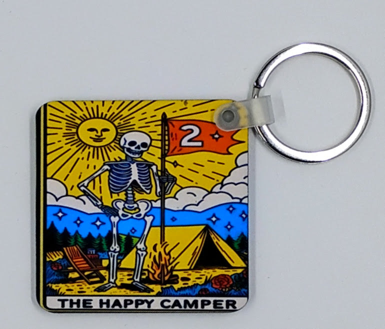 Handcrafted square keychain with a bright yellow sun and sky and a skeleton at a campsite and it says the happy camper
