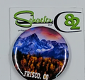 Small circular hat pin with a beautiful mountain scene and yellow tress in the foreground. Plus a pink sky