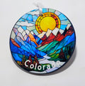 A circular ornament with a stained glass inspired mountain scenery and sun. 